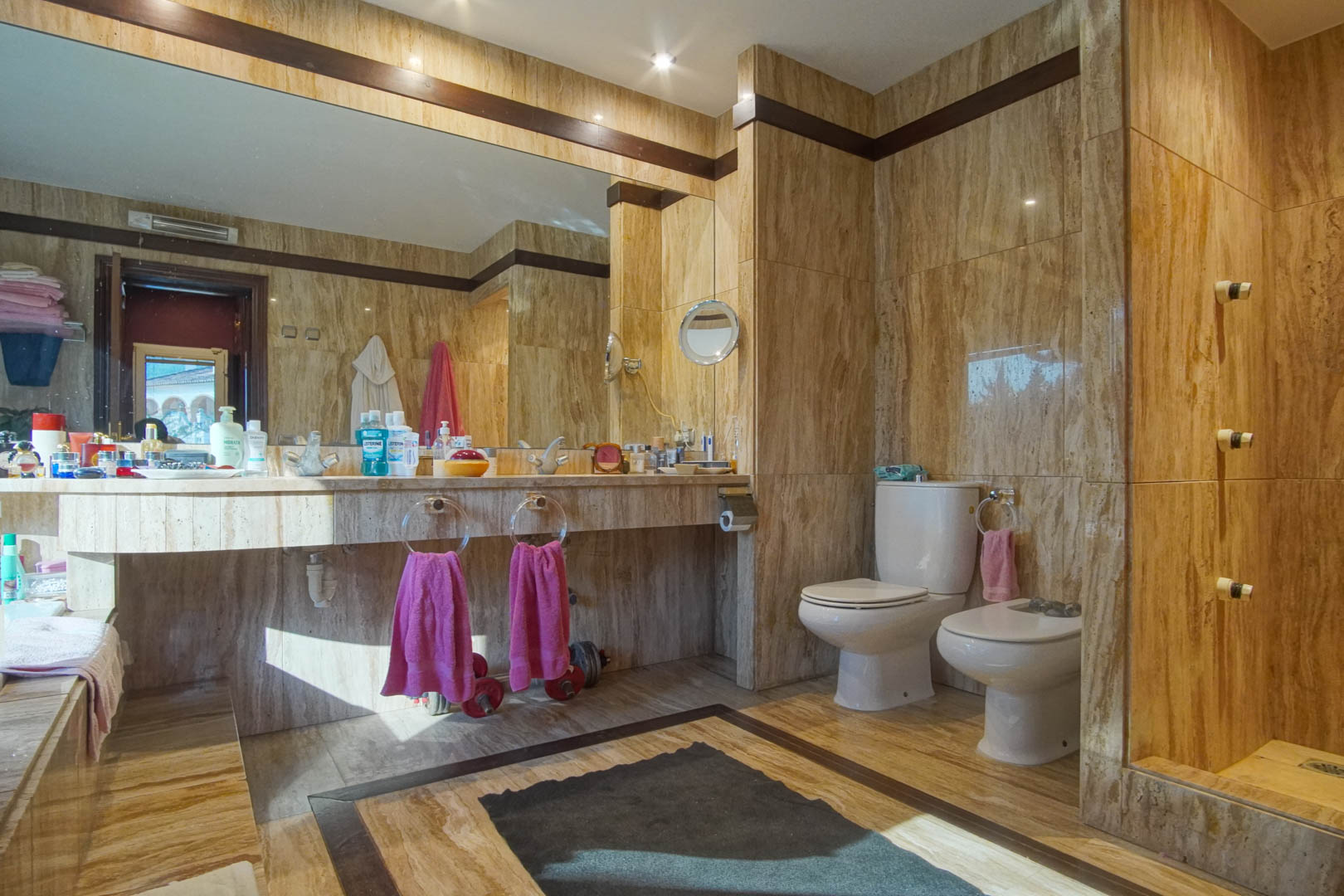 Bathroom fully clad in travertine marble featuring an integrated vanity, large wall mirror, recessed lighting, and matching bidet and toilet set.