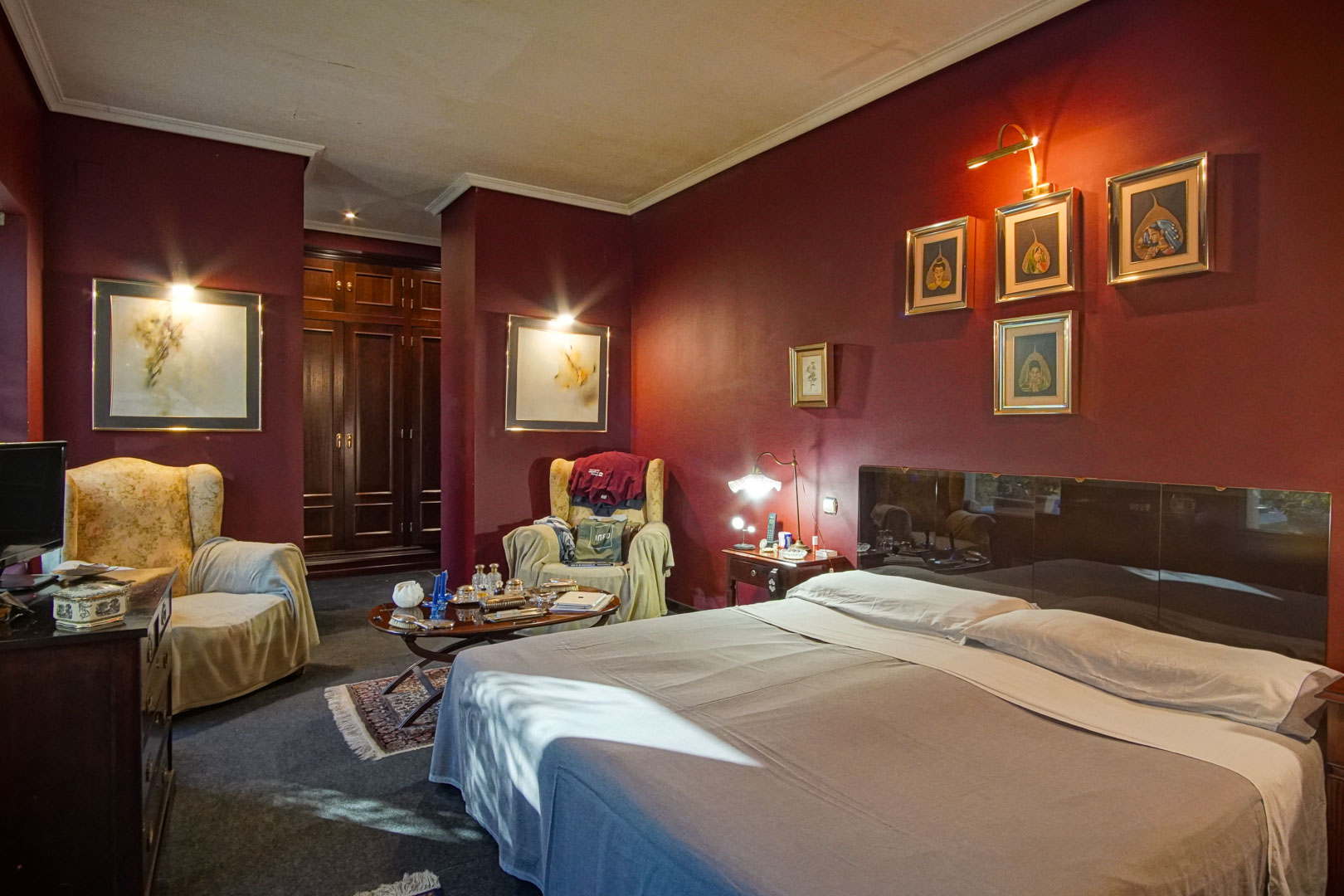 Bedroom featuring burgundy walls, a seating area with armchairs, and dark wood built-in wardrobes. Accent lighting highlights the artwork and the spacious layout.
