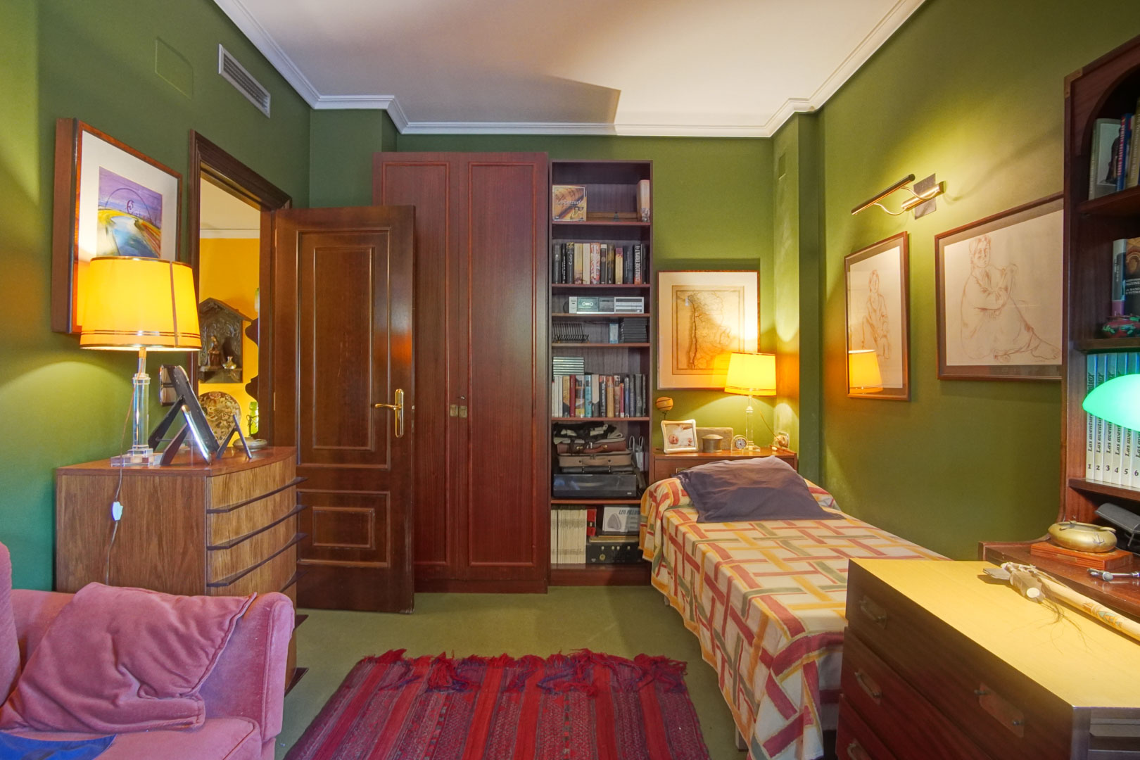 Bedroom featuring green walls, classic dark wood furniture, and a single bed. Includes a built-in bookshelf, warm ambient lighting, and ducted air conditioning.