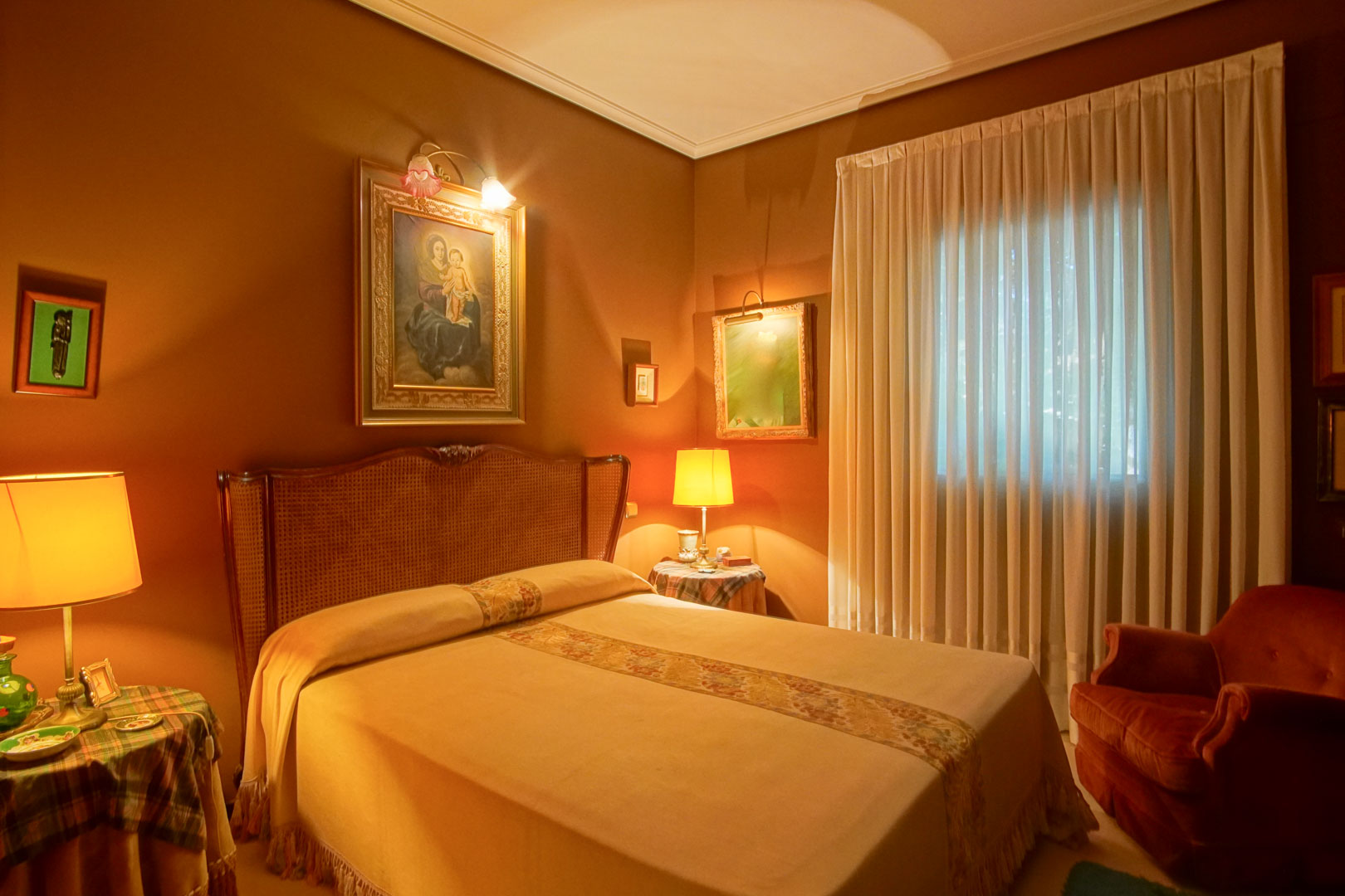 Classic bedroom featuring terracotta walls, a cane headboard, and warm ambient lighting. It includes framed artwork with dedicated lighting and a red velvet armchair.