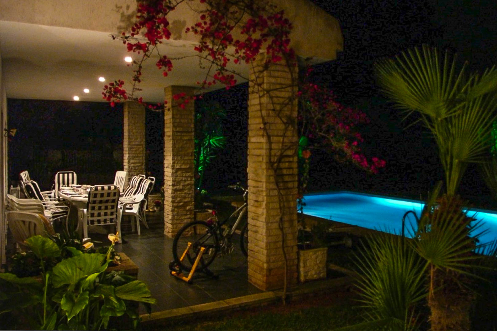 Covered terrace featuring exposed brick columns, outdoor dining area, and climbing bougainvillea next to an illuminated swimming pool at night.