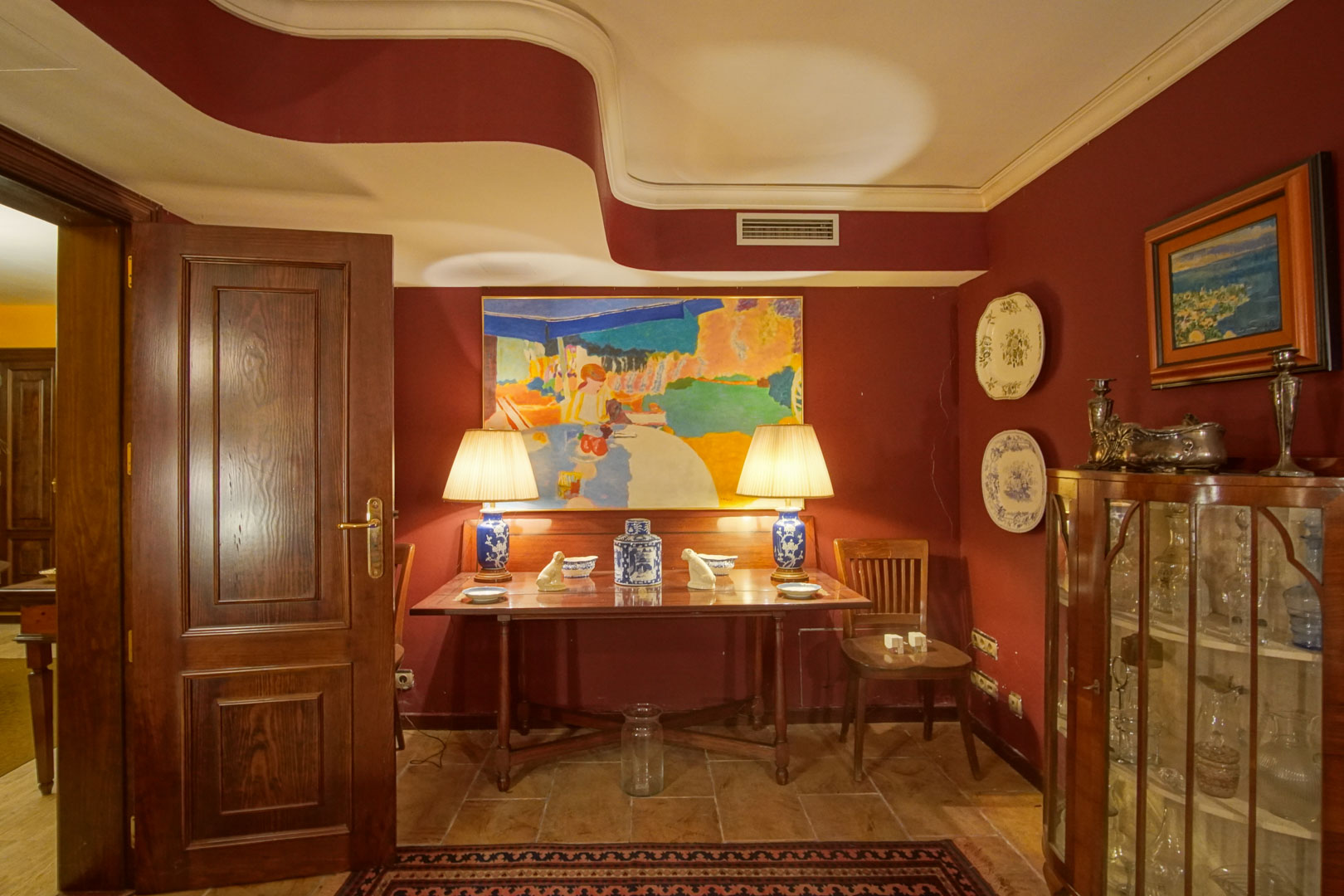 Elegant corner featuring burgundy walls, a unique wave-patterned ceiling, and classic wooden furniture. A glass display cabinet and ceramic lamps complement the natural stone flooring.