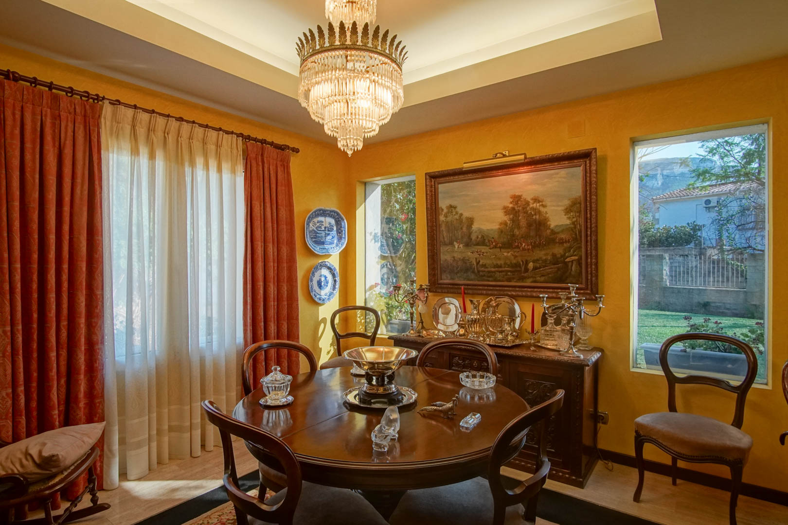 Elegant dining room featuring a round wooden table, crystal chandelier, and classic sideboard. The space includes ochre walls, tray ceiling with recessed lighting, and garden views.
