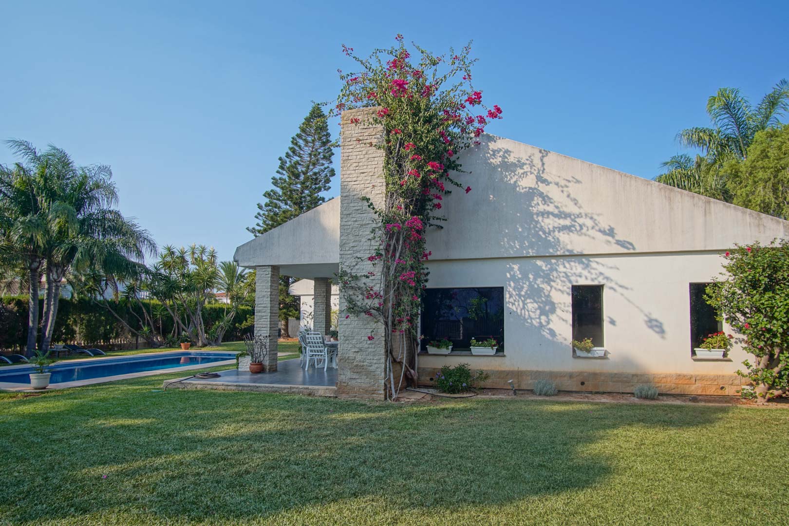 Exterior facade featuring a stone-clad chimney with climbing bougainvillea, a manicured lawn, and a private swimming pool in a Mediterranean setting.