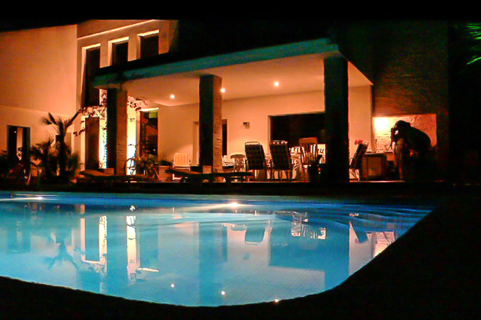 Illuminated covered terrace and private swimming pool at night, featuring stone-clad pillars and a spacious outdoor dining area.