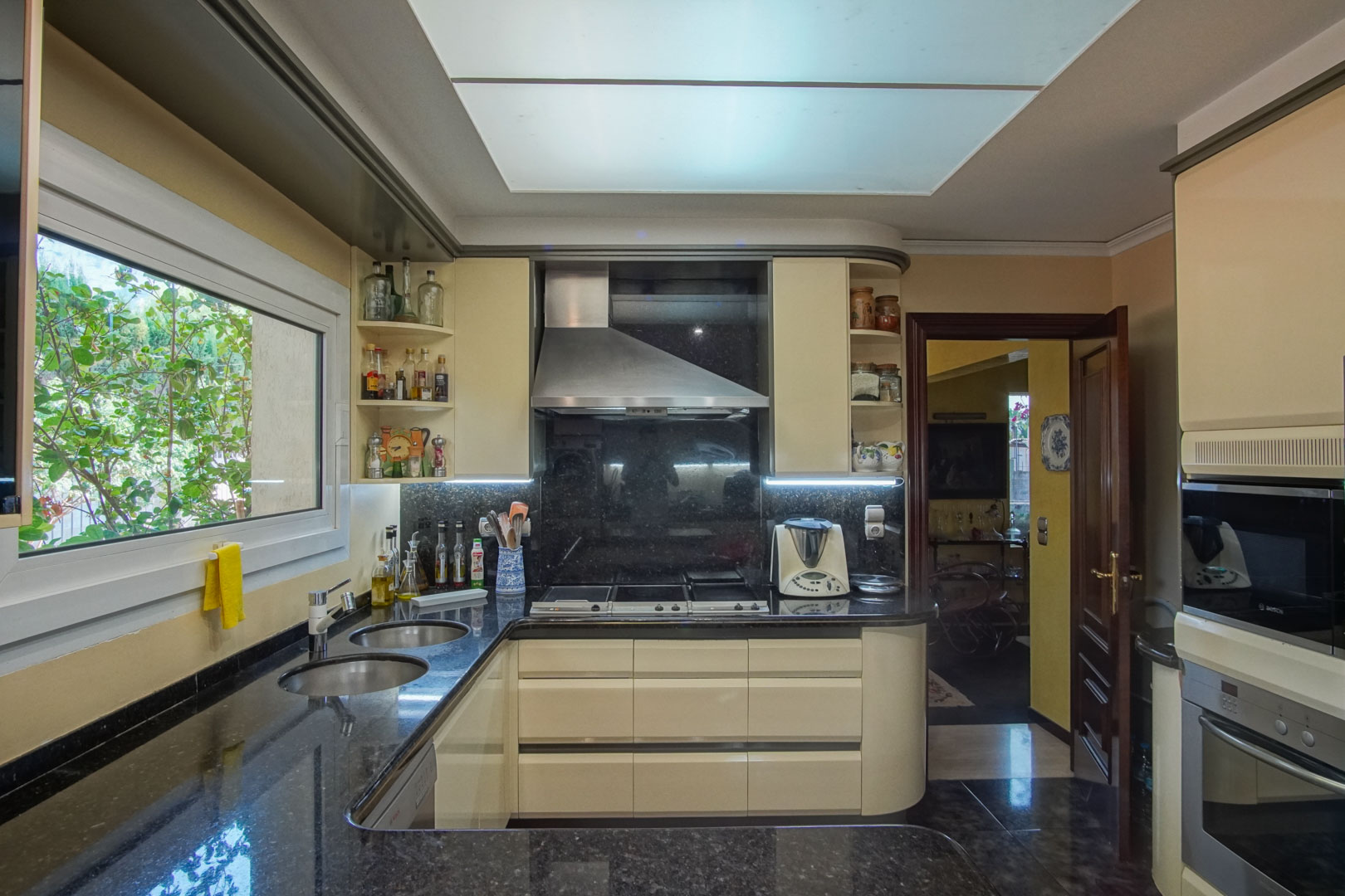 Independent kitchen featuring dark granite countertops, cream cabinetry, and integrated appliances, complemented by a large ceiling light panel and garden views.