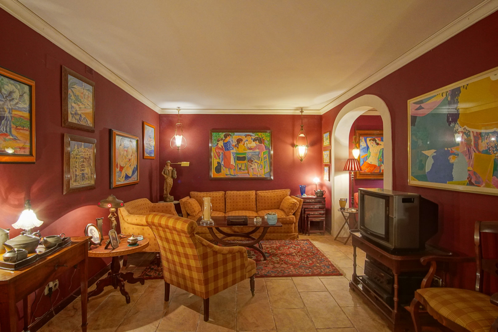 Living area featuring burgundy walls, rustic tile flooring, and classic upholstery. The space includes various artworks, warm ambient lighting, and a distinctive arched entrance.