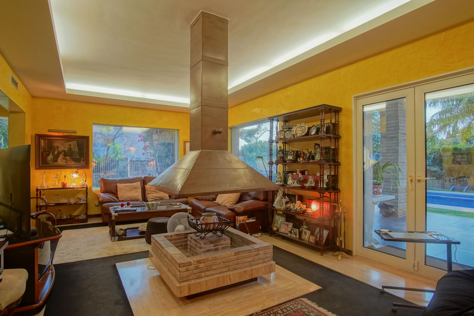 Living room featuring a central suspended fireplace with a brick base. Includes textured yellow walls, recessed ceiling lighting, and direct access to the pool area.