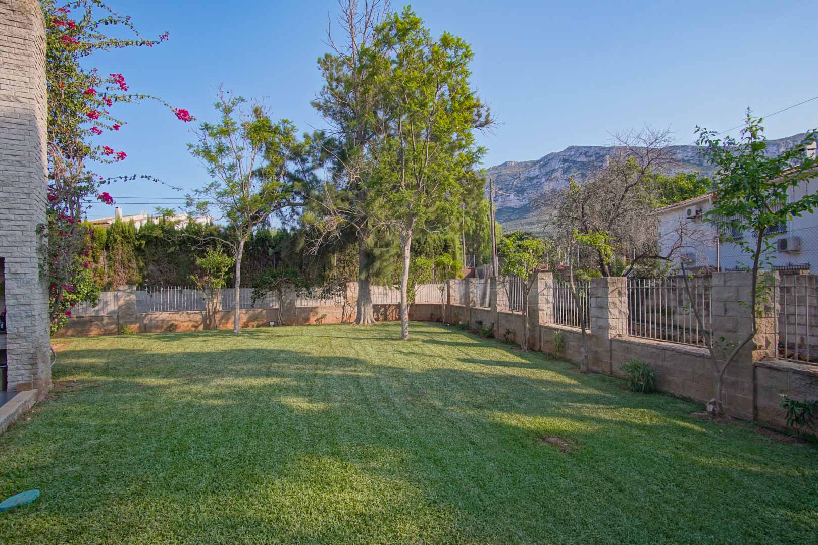 Manicured natural lawn surrounded by a stone and metal perimeter fence, featuring mature trees and a direct view of the Montgó mountain.