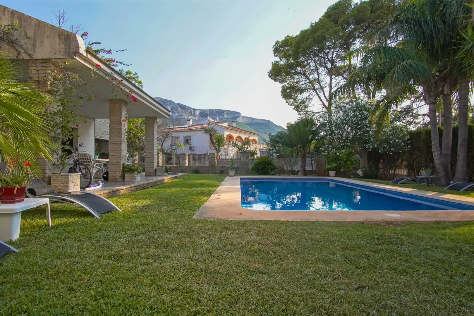 Mediterranean garden featuring a natural lawn, rectangular pool, and covered terrace with stone pillars. The Montgó mountain provides a scenic backdrop in the distance.
