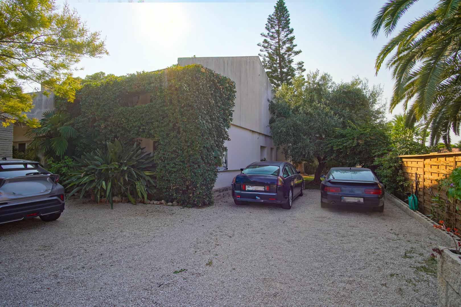 Outdoor parking area featuring a gravel surface, surrounded by mature Mediterranean vegetation and a wooden perimeter fence. Space accommodates multiple vehicles.