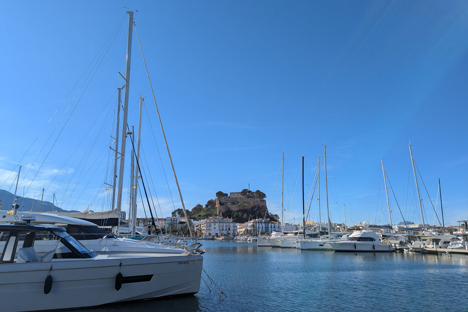 Panoramic view of Dénia's marina featuring luxury yachts and the historic castle overlooking the harbor under a clear blue sky.