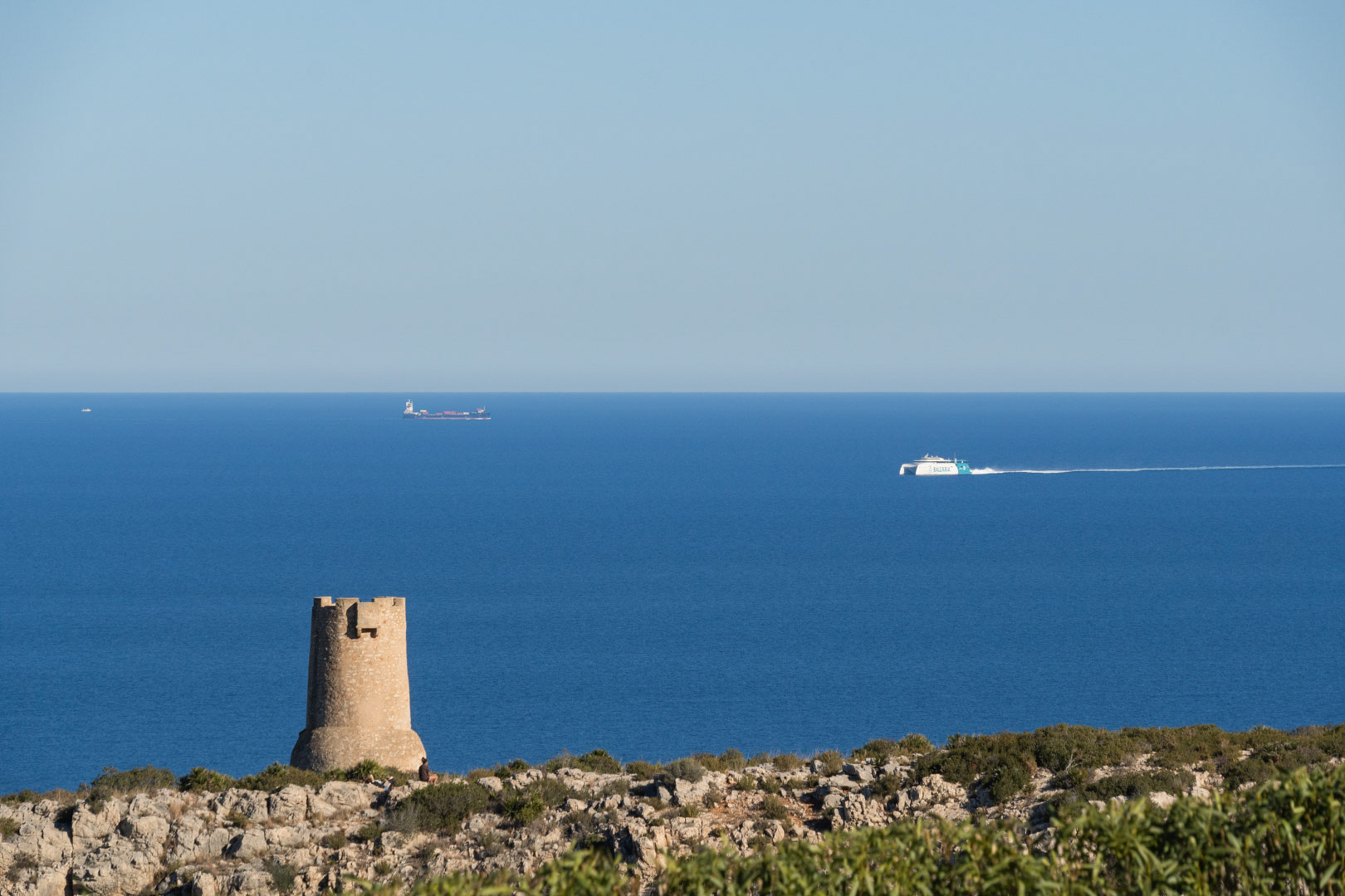 Panoramic views of the Dénia coastline featuring the historic Torre del Gerro watchtower and ferries crossing the Mediterranean Sea toward the Balearic Islands.