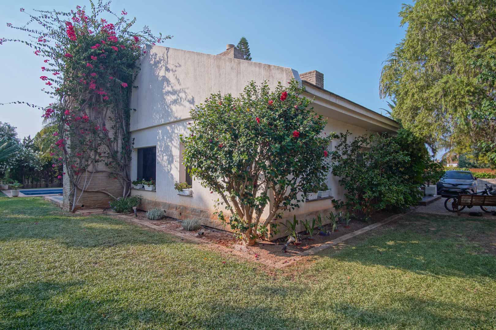Side facade of the villa featuring a natural lawn, climbing bougainvillea on a stone wall, and Mediterranean shrubs under a clear blue sky.
