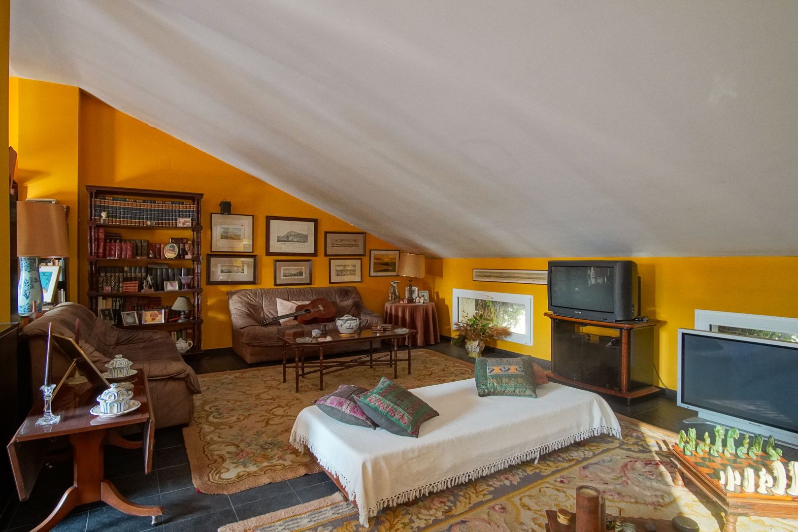 Spacious attic featuring sloped ceilings, mustard yellow walls, and dark tiling. This versatile living area includes bookshelves, seating, and natural light.