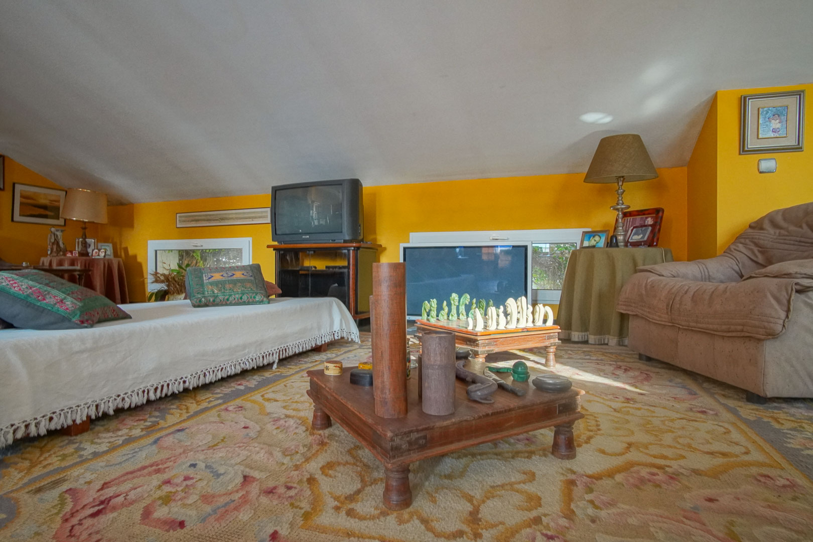 Spacious attic room with slanted ceilings, mustard yellow walls, and a leisure area. Features patterned carpeting, low wooden tables, and natural light through floor-level windows.