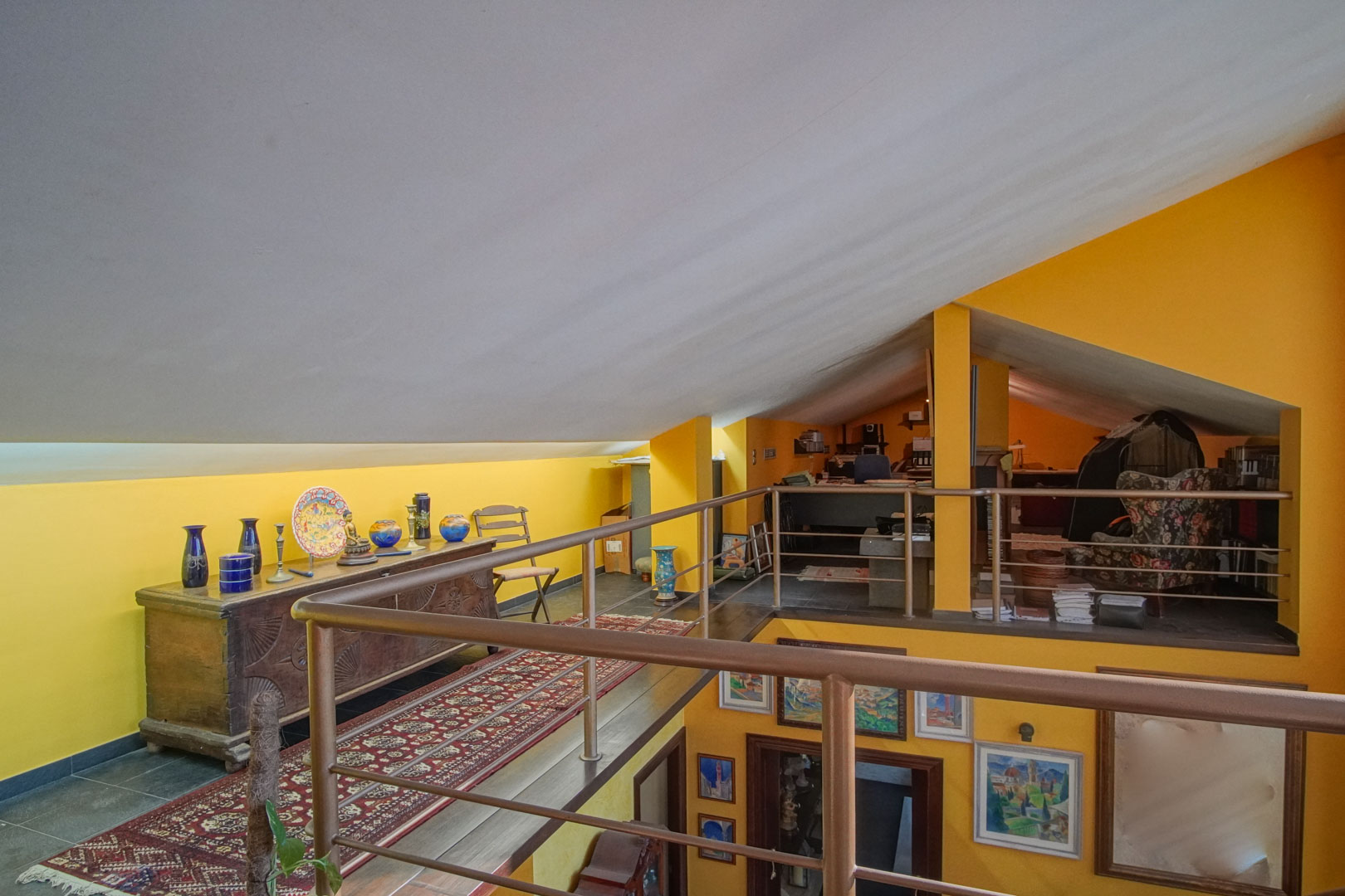 Spacious mezzanine attic featuring sloped ceilings, vibrant yellow walls, and metal railings. The area includes a decorative wooden sideboard and overlooks the lower living space.