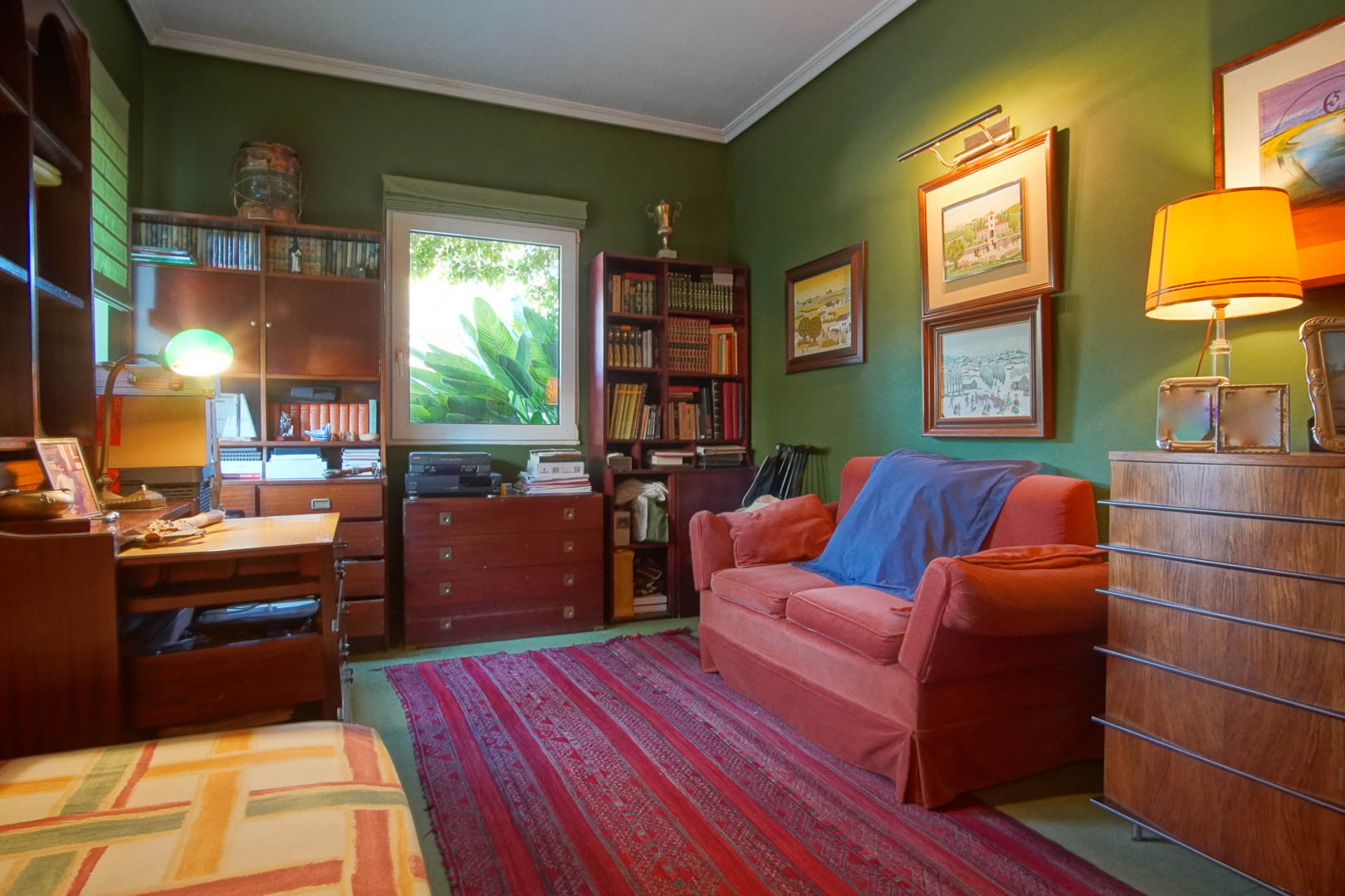Study with green walls and classic wooden furniture. It features bookshelves, a desk, a red sofa, and natural light with views of the outdoor greenery.