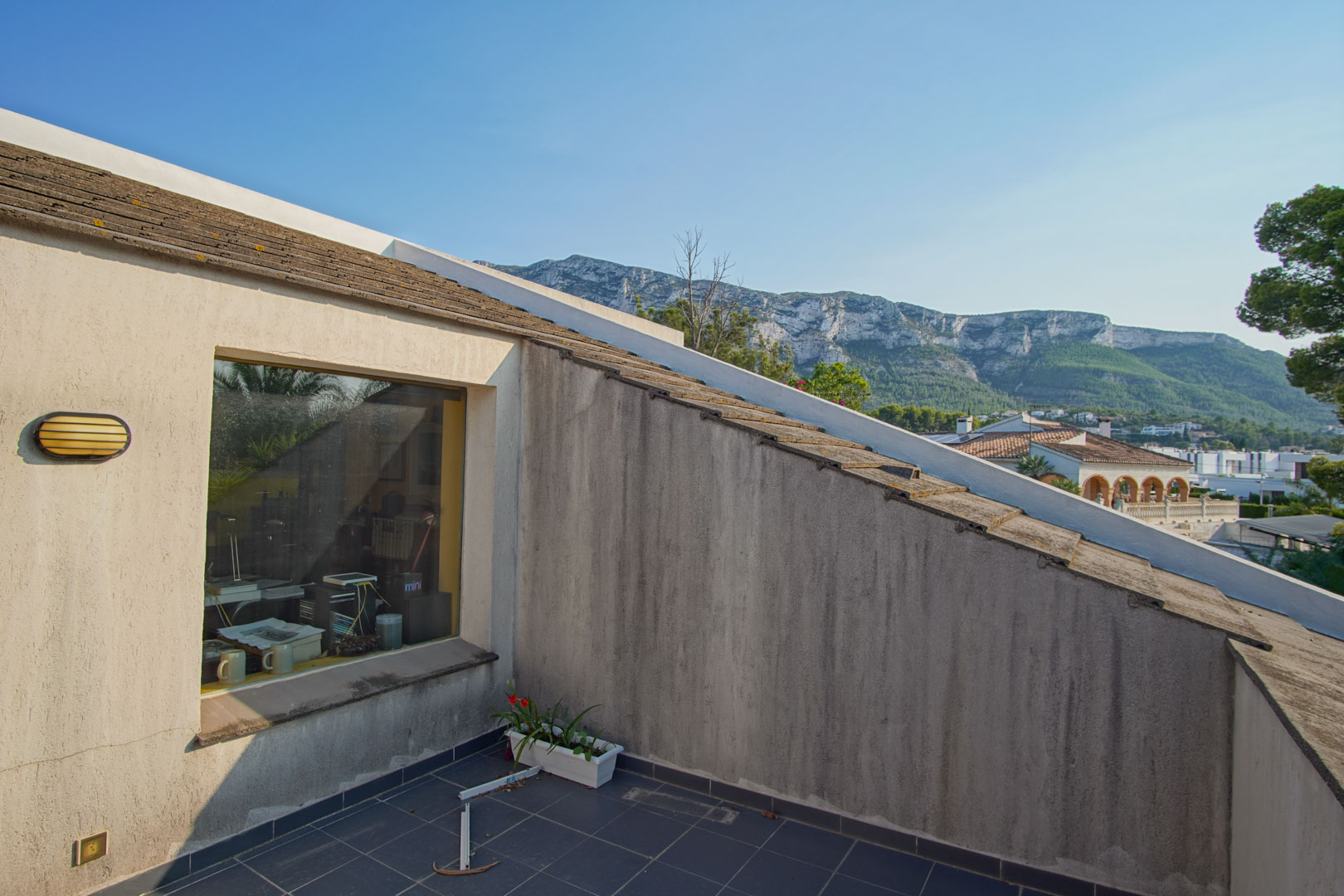 Upper-floor balcony featuring dark ceramic tiling and direct views of the Montgó mountain. A large window connects the exterior space with the interior office area.