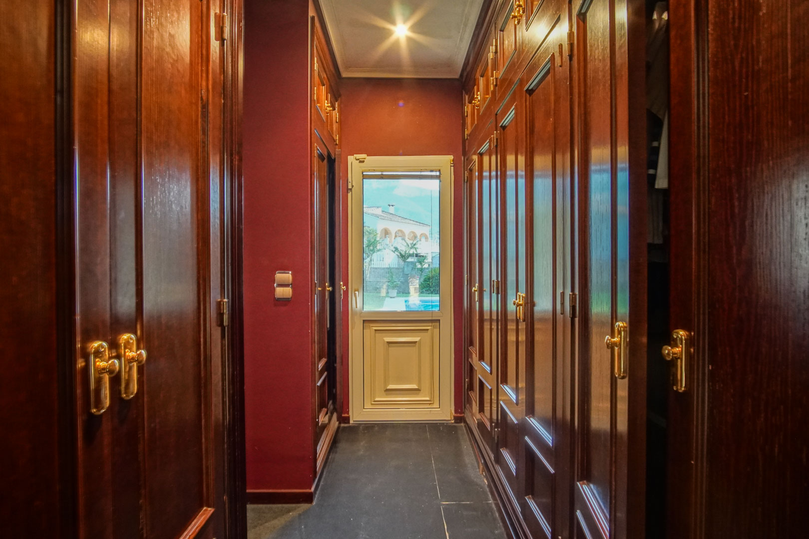 Walk-in closet with dark wood built-in wardrobes, gold hardware, burgundy walls, and a glass door leading to the exterior.