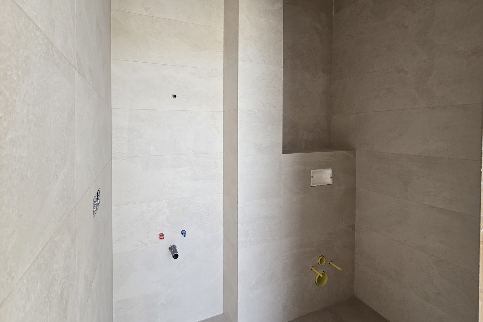 Bathroom under construction featuring sand-toned porcelain wall tiles, plumbing for a vanity, and a built-in cistern for a wall-mounted toilet with a recessed niche.