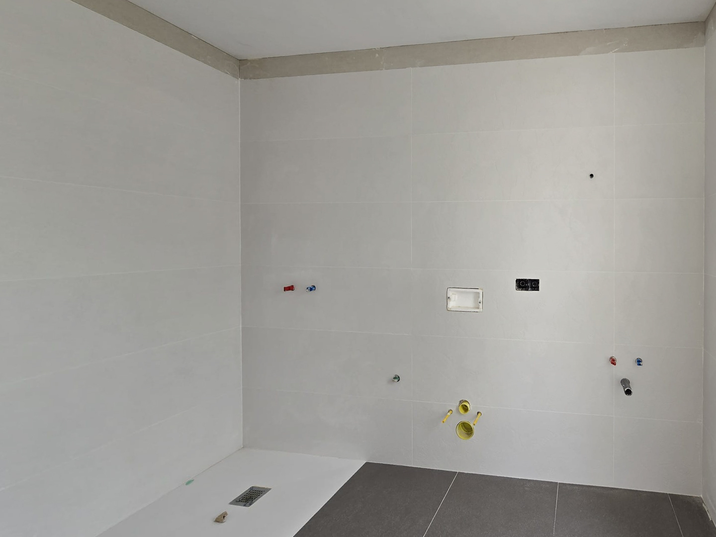Bathroom under construction showing large white wall tiles, a floor-level shower tray, and prepared plumbing and electrical points for the vanity and toilet.