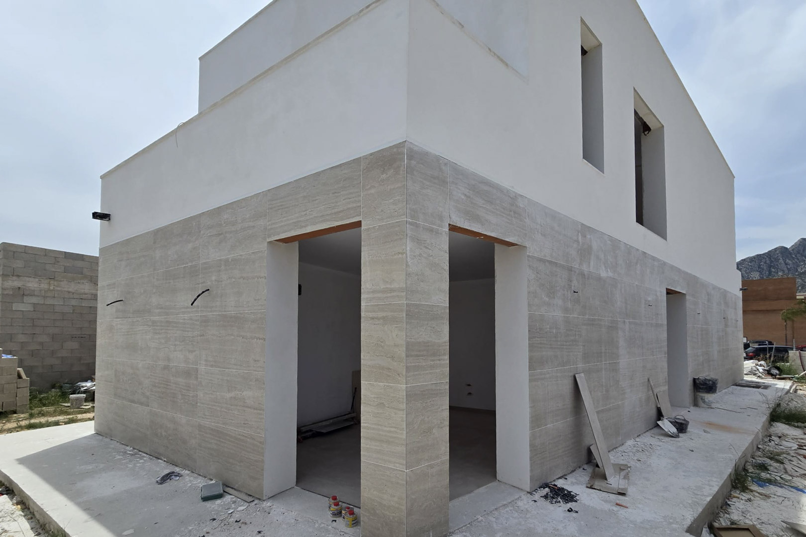 Exterior facade under construction featuring stone-effect cladding on the ground floor and white finishes above, showcasing modern geometric lines and large openings.