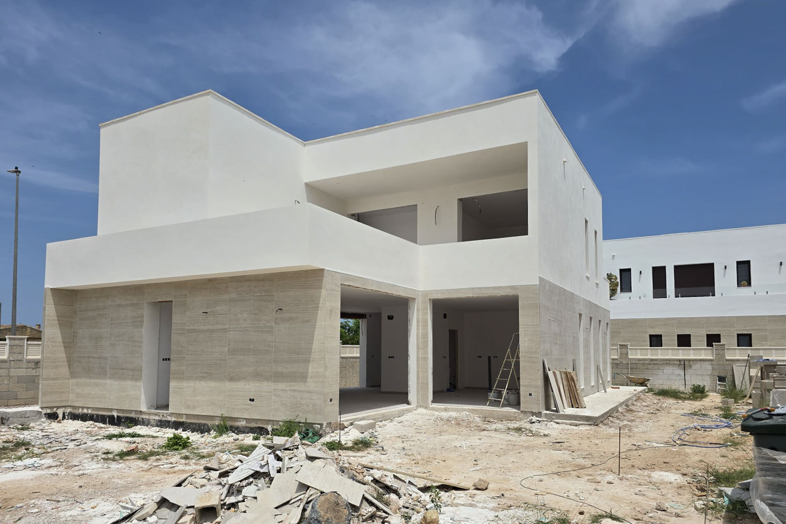Exterior view of a modern villa under construction featuring stone-clad ground floor walls, white upper volumes, and large structural openings for floor-to-ceiling windows.