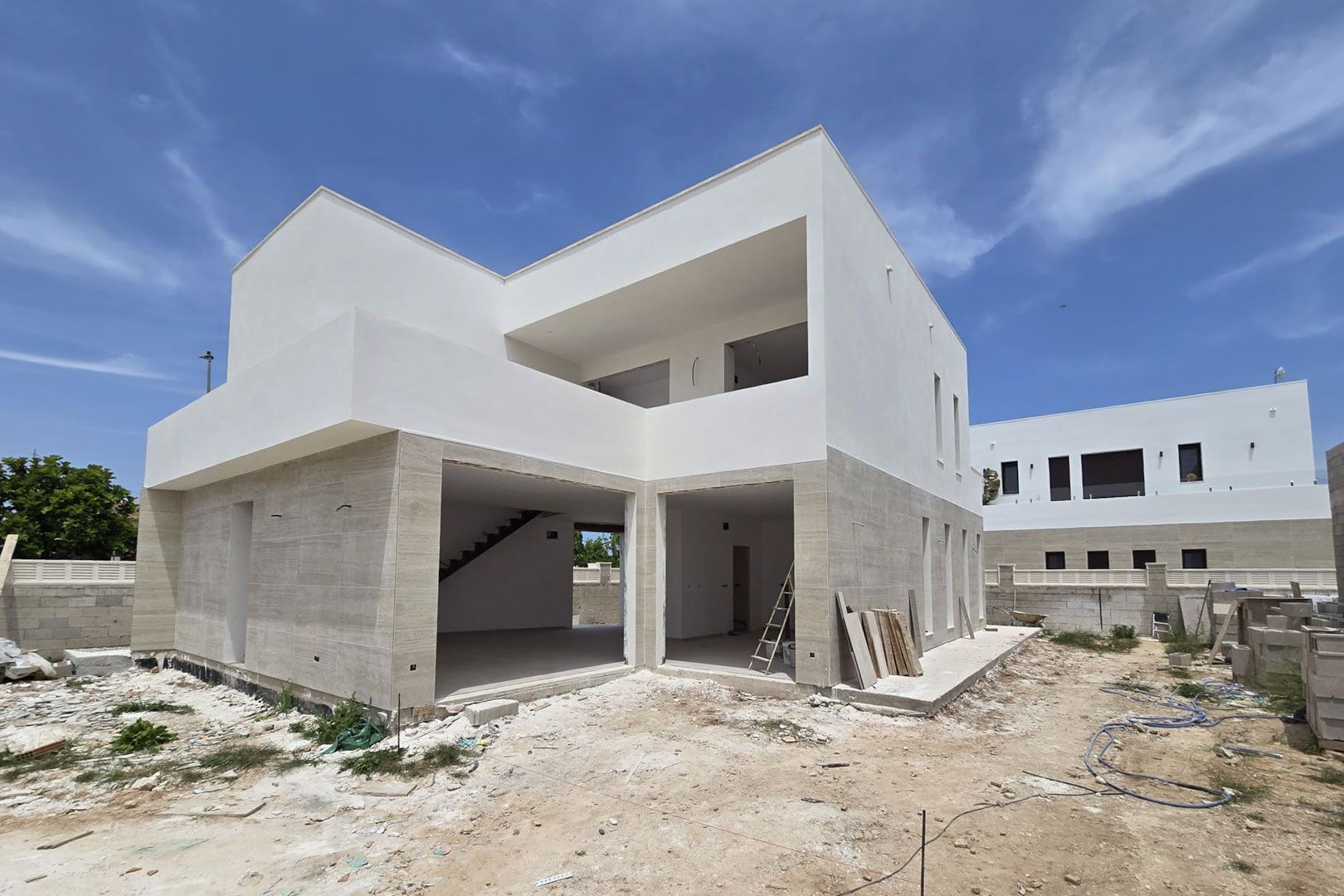Exterior view of the villa under construction, featuring stone cladding on the ground floor, white upper volumes, and large openings for floor-to-ceiling windows.
