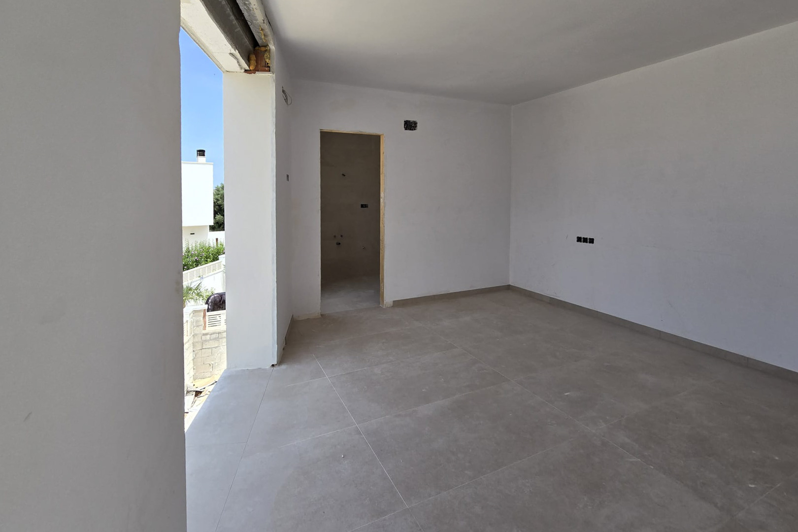 Interior room under construction featuring large-format grey floor tiles, white walls, and direct outdoor access. An ensuite bathroom entrance and electrical points are visible.