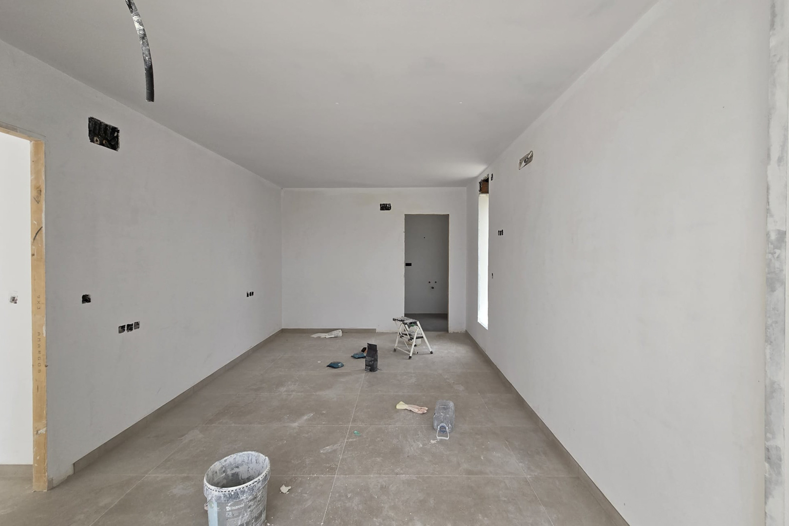 Interior view under construction featuring large-format porcelain floor tiles and smooth white walls. Electrical pre-installations and doorways to adjacent rooms are visible.