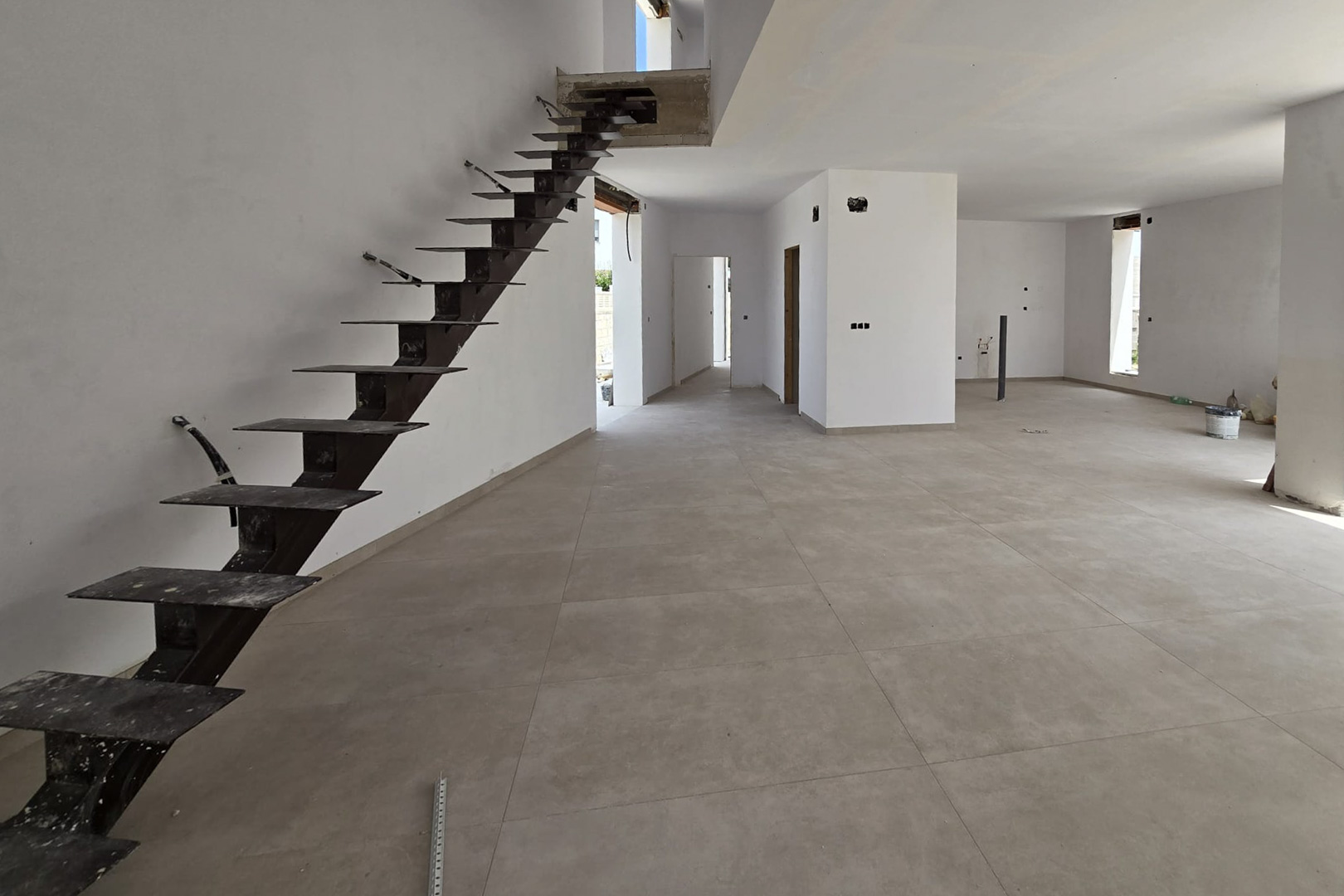 Open-plan ground floor under construction featuring large-format grey porcelain tiles and a cantilevered metal staircase leading to the upper floor.