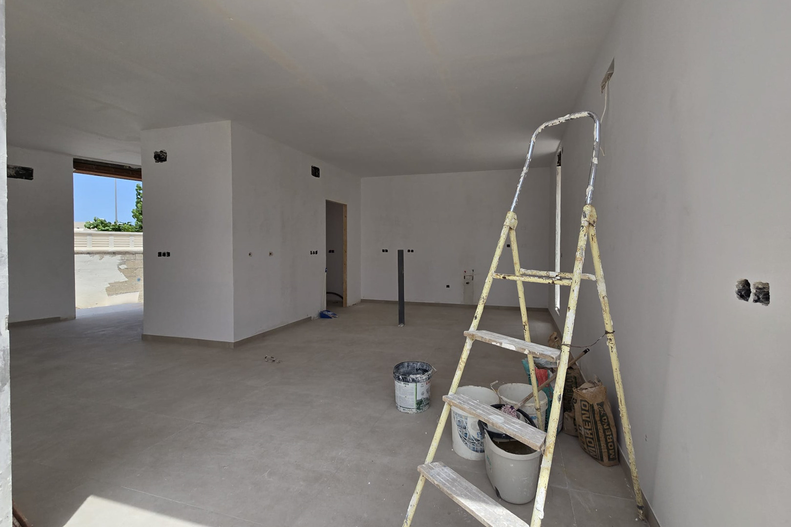 Open-plan interior in the finishing phase with large-format ceramic flooring and white walls. Electrical pre-installations and access to the exterior are visible.