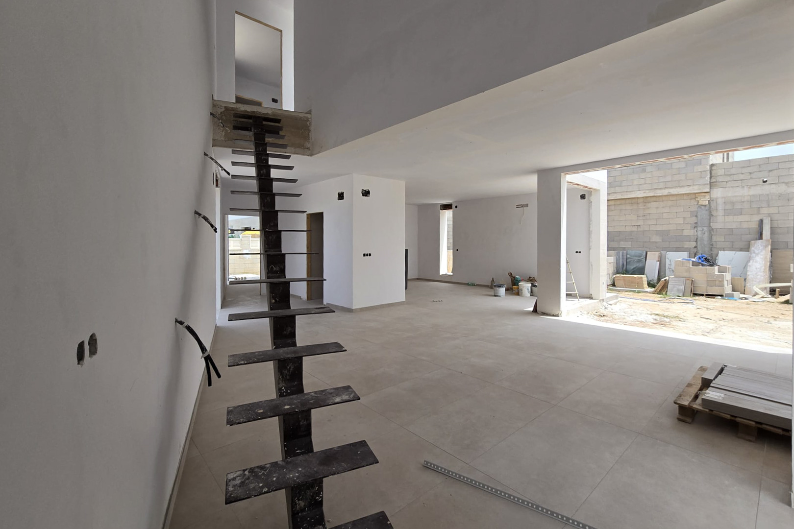 Open-plan interior under construction featuring a sleek metal staircase, large-format porcelain flooring, and a double-height ceiling for a spacious feel.