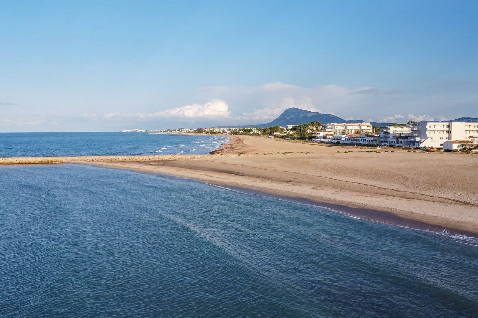 Panoramic view of the wide sandy beach near the property, featuring a stone breakwater, the Mediterranean Sea, and the Montgó mountain silhouette.