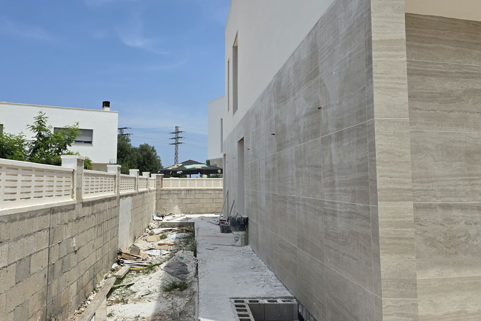 Side facade under construction featuring large-format cream stone-effect cladding, highlighting the modern architectural lines and the perimeter boundary wall.
