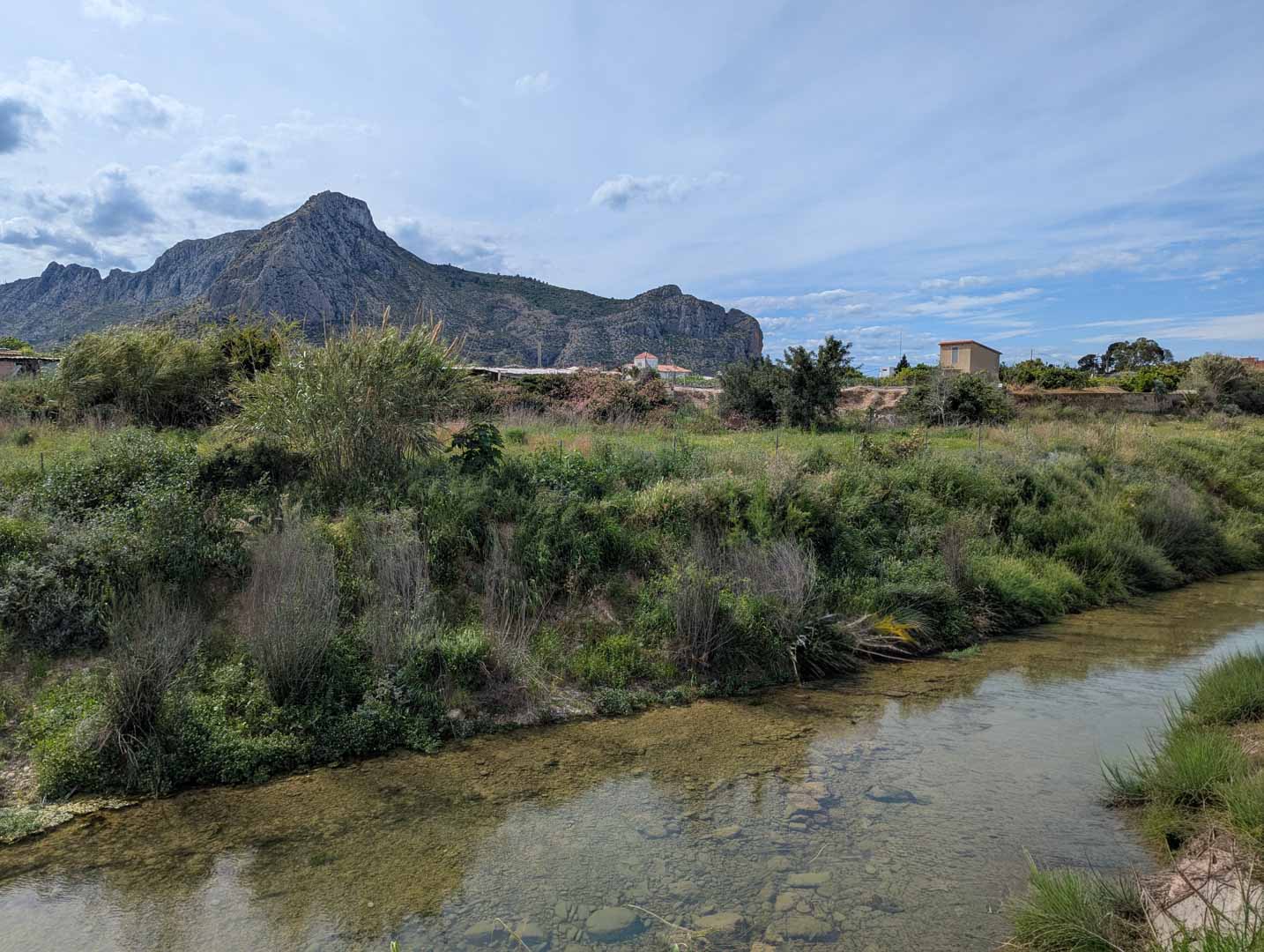 View of the natural surroundings featuring a clear stream with a rocky bed and lush vegetation, set against a backdrop of rugged mountain peaks.