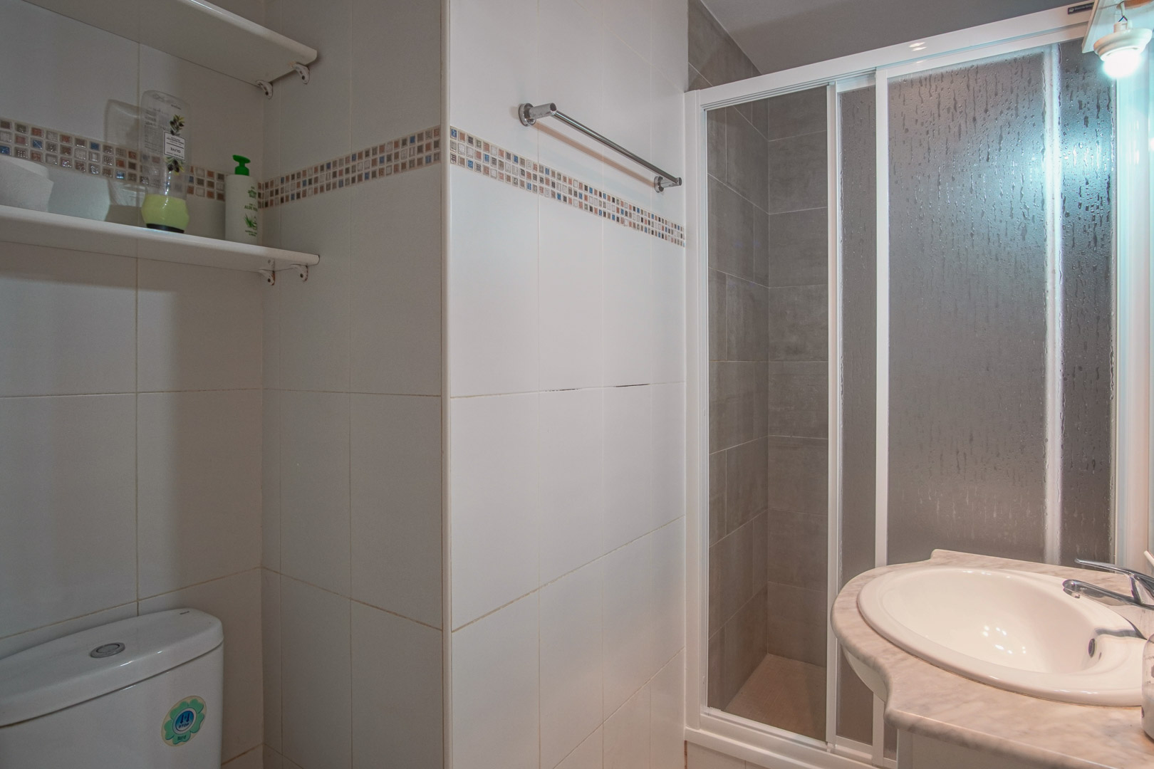 Bathroom featuring white tiled walls with decorative borders, a walk-in shower with sliding frosted doors, and a marble-top vanity with integrated sink.