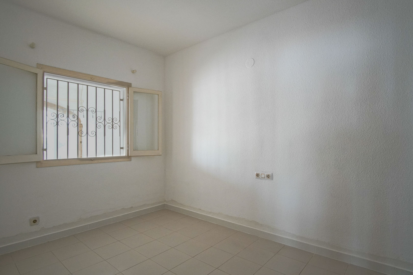 Bright bedroom featuring white textured walls, light ceramic tile flooring, and an exterior window equipped with decorative security bars.