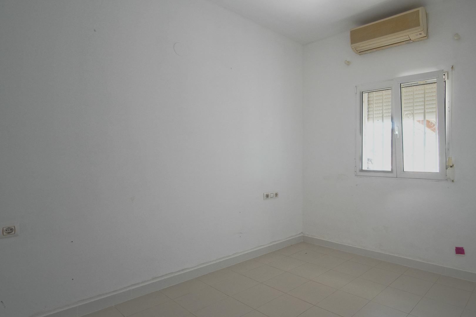 Bright bedroom featuring white walls, tiled flooring, and a wall-mounted air conditioning unit. Includes a PVC window with integrated shutters and power outlets.