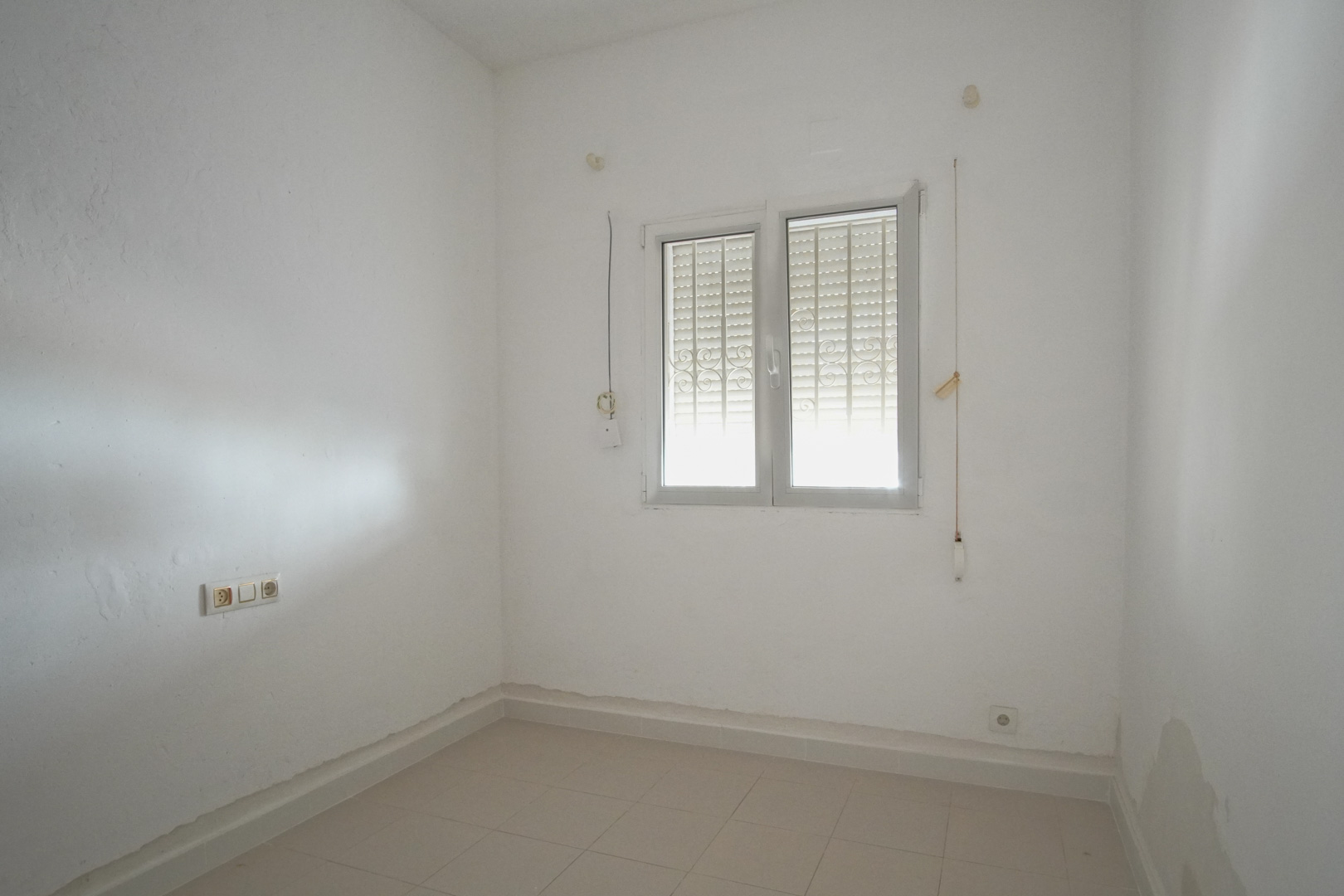 Bright room featuring white walls, ceramic tile flooring, and a double-pane window equipped with security bars and integrated shutters.