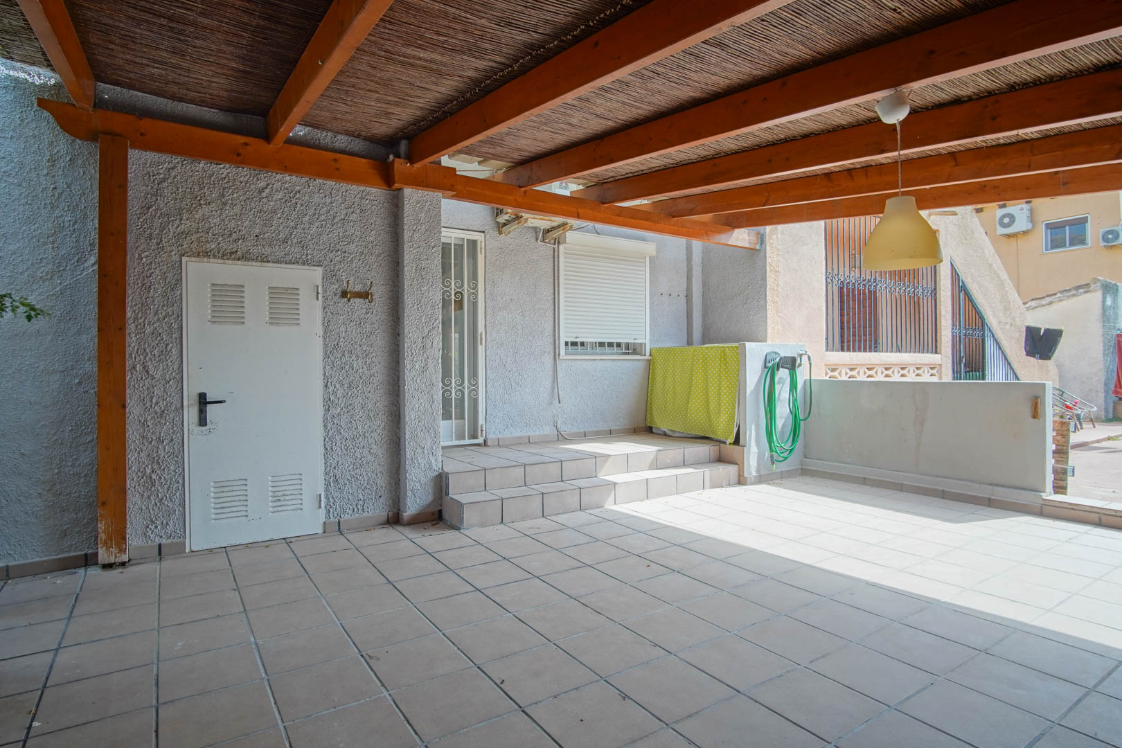 Covered outdoor terrace featuring a wooden pergola with reed roofing, tiled flooring, and direct access to the interior via steps and a white security door.