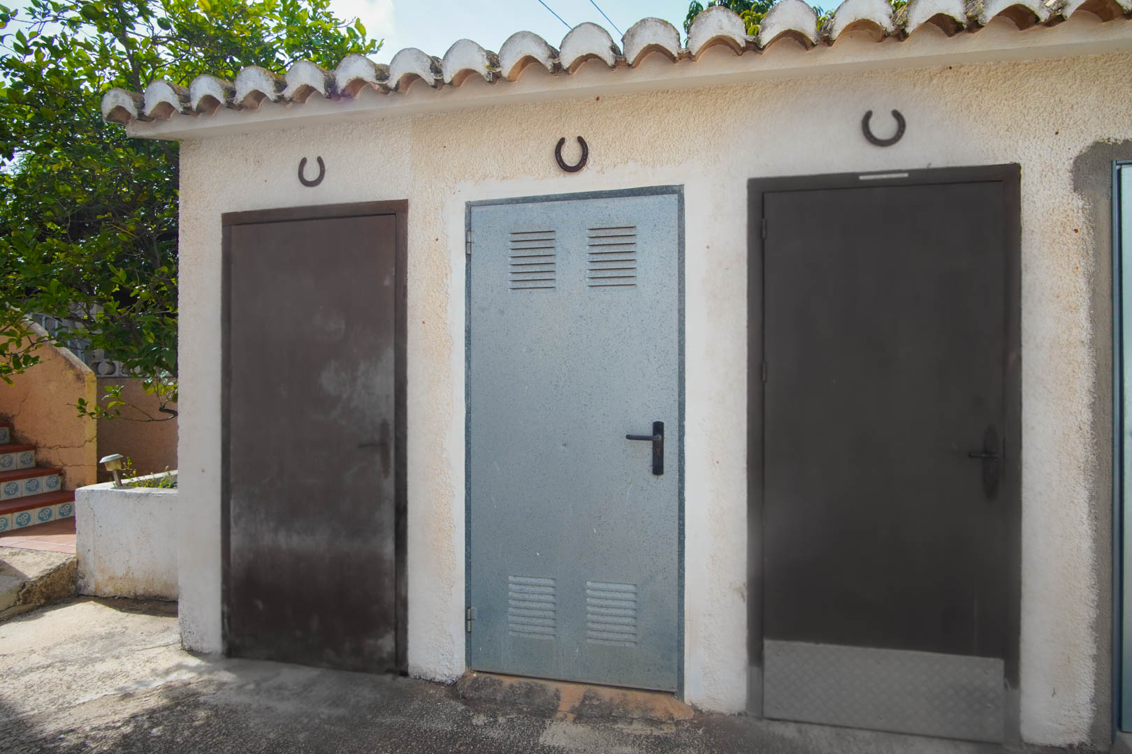 Exterior storage area featuring three metal utility doors under a traditional tiled roof, accented with decorative horseshoes on a textured white wall.