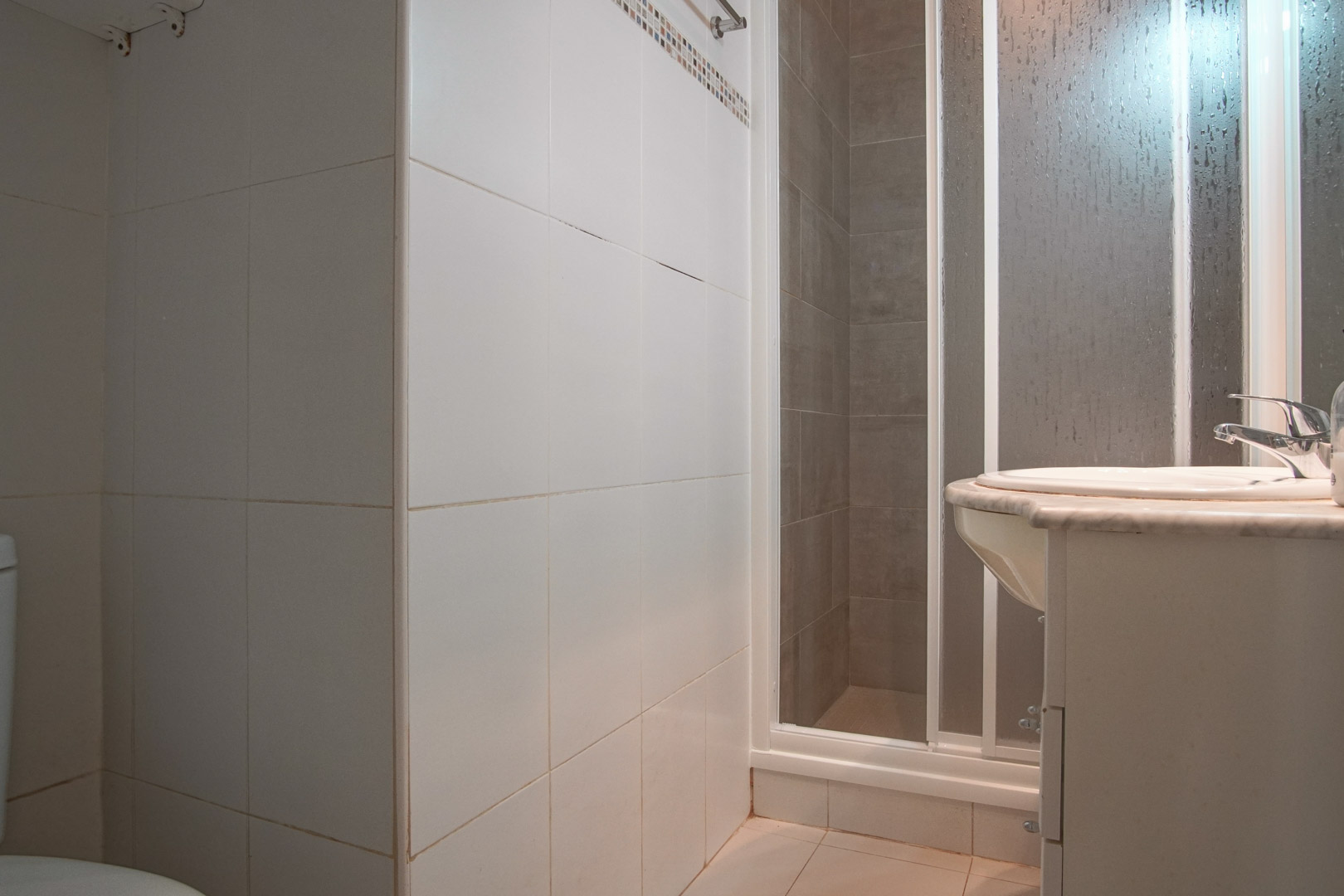 Functional bathroom featuring white tiled walls, a shower cabin with a frosted screen, and a washbasin set into a marble countertop.