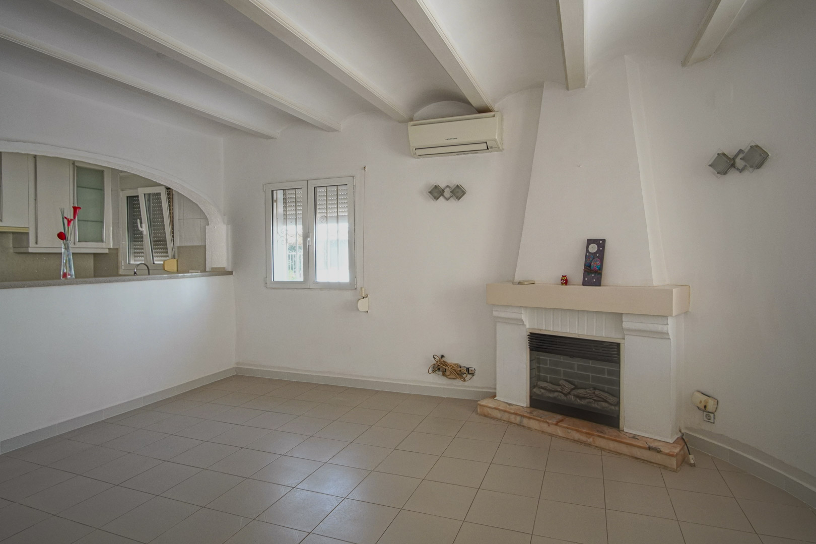 Living area featuring a built-in fireplace, white beamed ceilings, and split air conditioning. An arched opening connects the space to the kitchen.