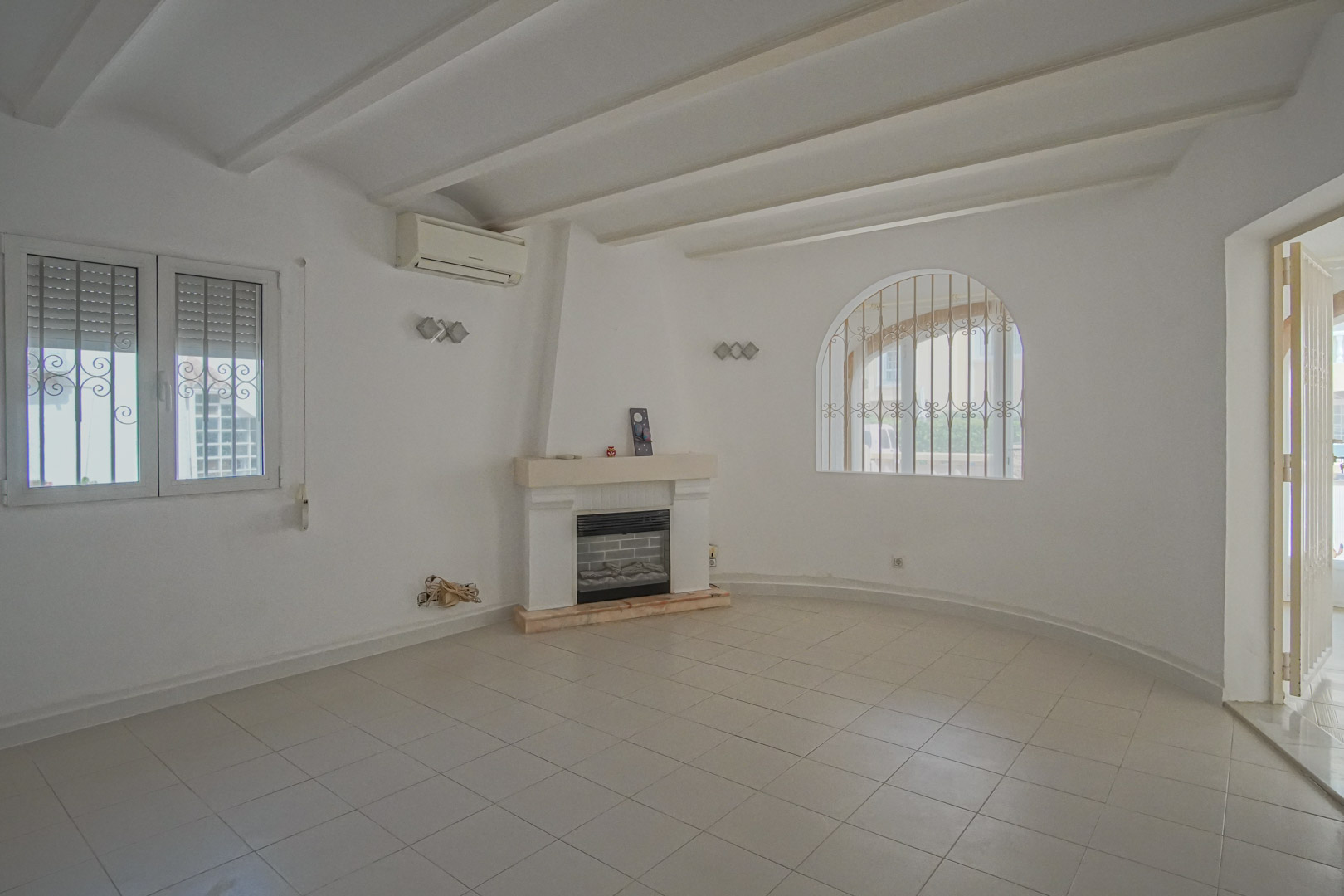 Living room featuring a central fireplace, white exposed beams, and an arched window with decorative security bars. Equipped with air conditioning and tiled flooring.