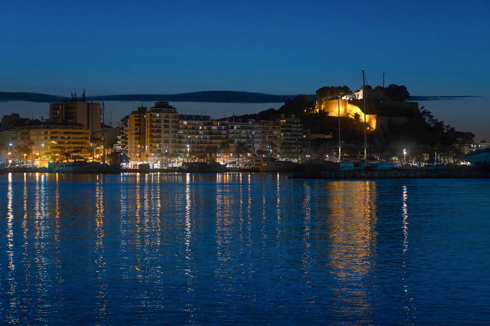 Night view of Dénia's harbor showcasing the illuminated historic castle and the city skyline reflected on the calm Mediterranean sea.