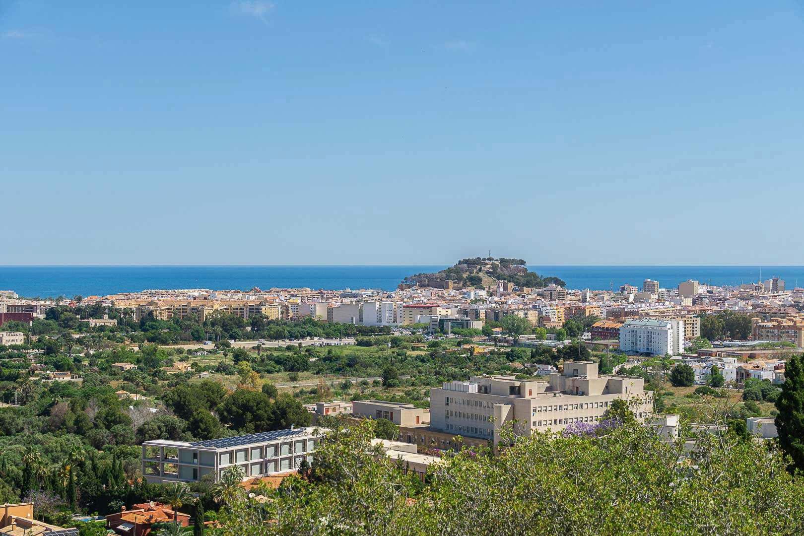 Panoramic view of Dénia featuring the historic castle on the hill, the Mediterranean Sea horizon, and the urban landscape integrated with green areas.