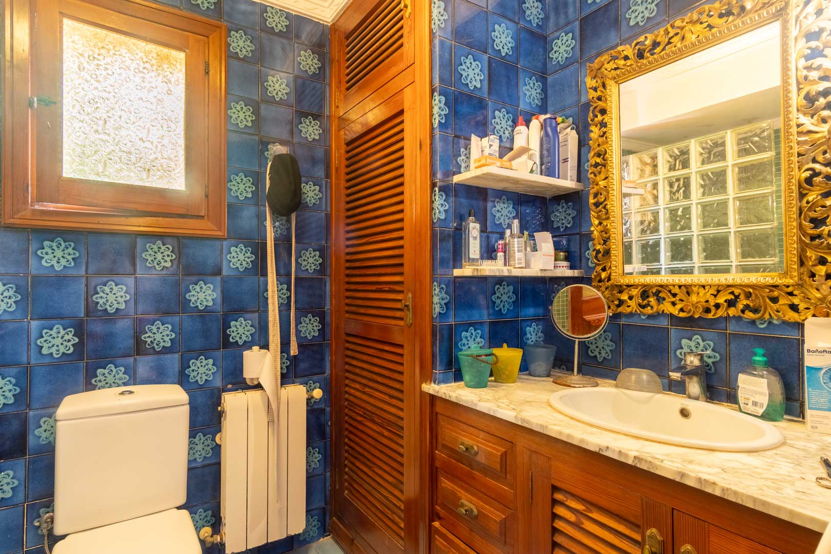 Bathroom featuring blue patterned tiles, a wooden vanity with a marble top, and a classic ornate gold-framed mirror complemented by natural wood accents.