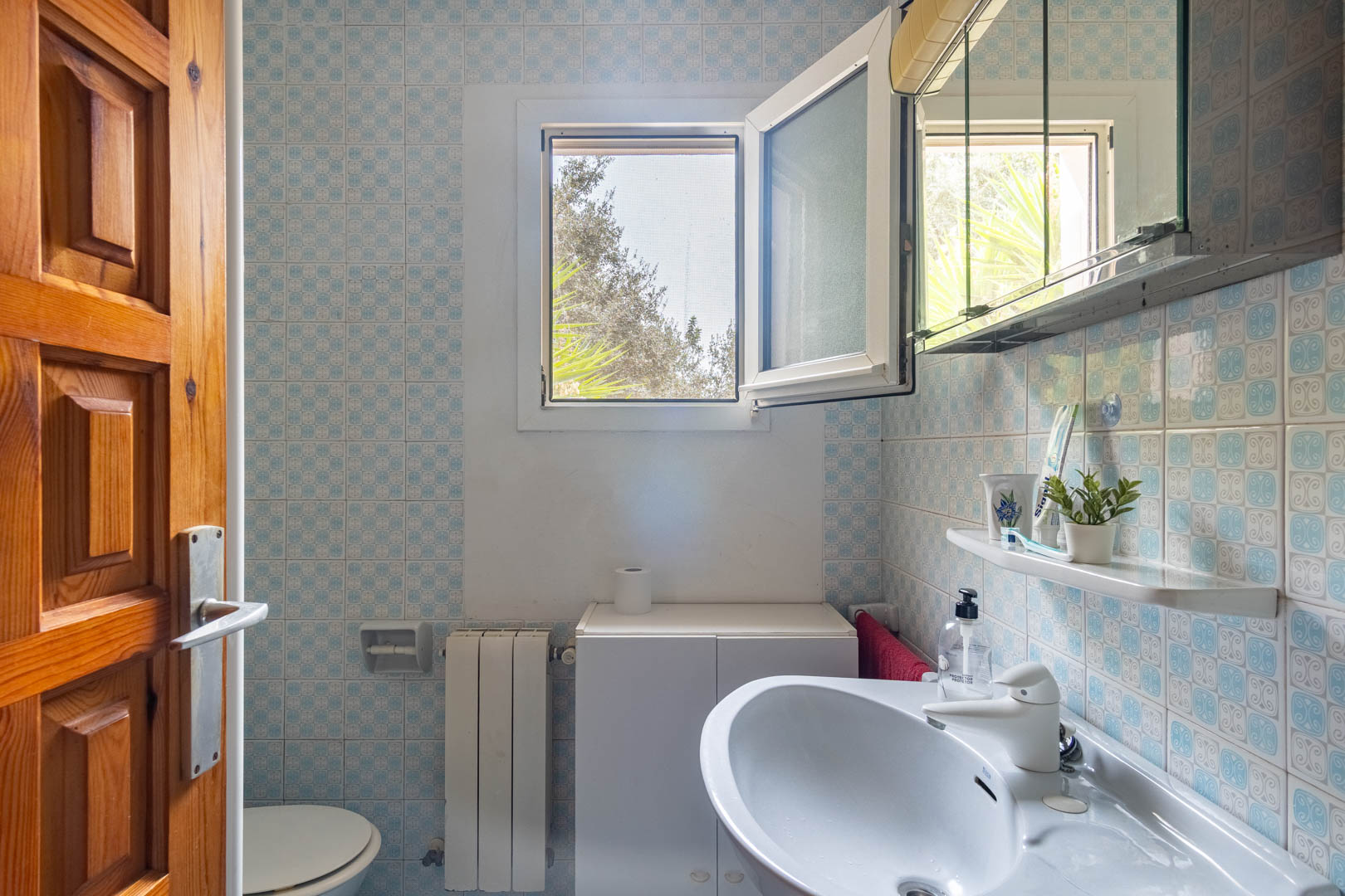 Bathroom featuring blue patterned wall tiles, a white ceramic sink, and a window with a fly screen providing natural light and ventilation.